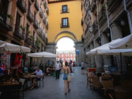 Kobieta spacerująca po wąskiej uliczce z kawiarniami i parasolami w Madrycie, Hiszpania, prowadzącej do łuku z widokiem na Plaza Mayor, otoczona zabytkowymi budynkami.