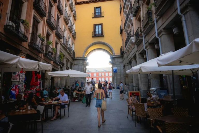 Kobieta spacerująca po wąskiej uliczce z kawiarniami i parasolami w Madrycie, Hiszpania, prowadzącej do łuku z widokiem na Plaza Mayor, otoczona zabytkowymi budynkami.