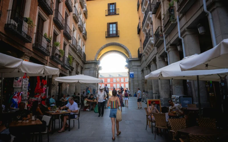 Kobieta spacerująca po wąskiej uliczce z kawiarniami i parasolami w Madrycie, Hiszpania, prowadzącej do łuku z widokiem na Plaza Mayor, otoczona zabytkowymi budynkami.