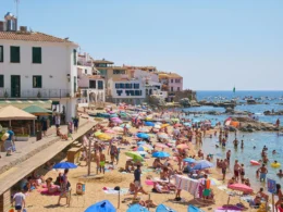 Zatłoczona plaża w nadmorskim miasteczku na Costa Brava, Hiszpania, z kolorowymi parasolami, turystami kąpiącymi się w morzu oraz białymi budynkami w tle.