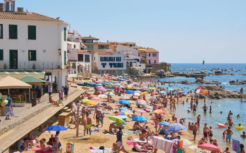 Zatłoczona plaża w nadmorskim miasteczku na Costa Brava, Hiszpania, z kolorowymi parasolami, turystami kąpiącymi się w morzu oraz białymi budynkami w tle.